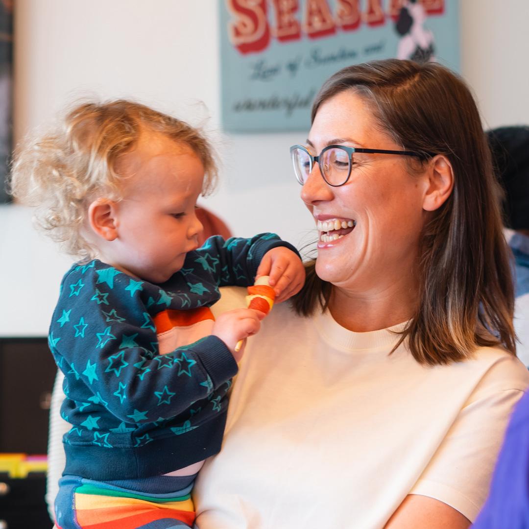 A women is holding a baby and smiling widely, in a family centre setting.