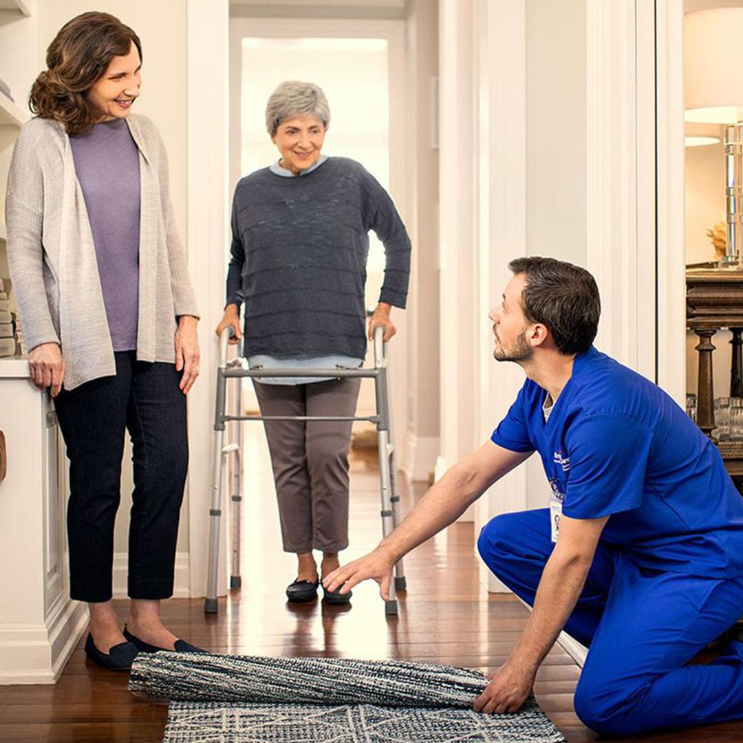 A nurse moves a crumpled rug at the home of an elderly couple, who stand smiling in the doorway of a hallway.