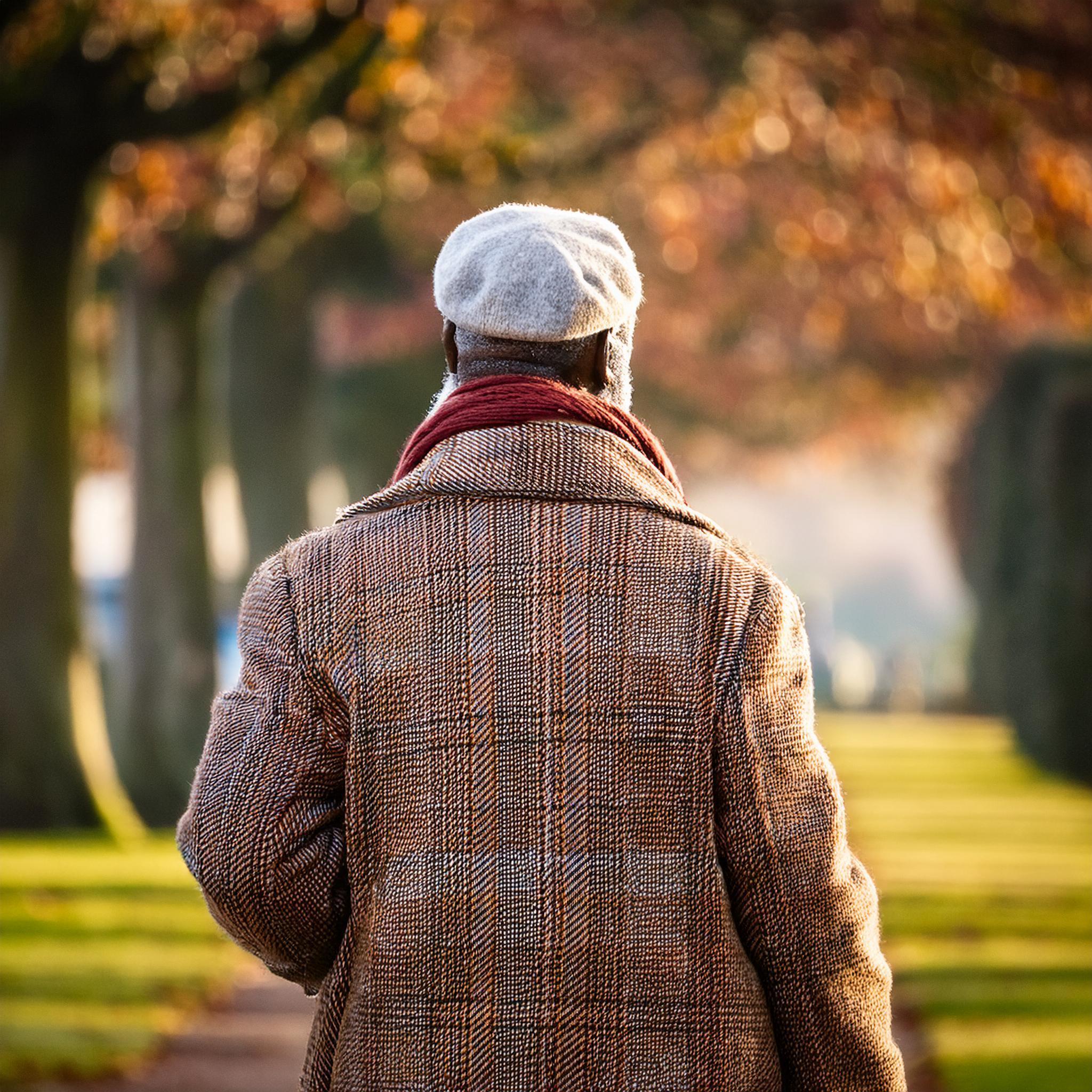 An elderly man wearing a winter coat and flat cap walking through a park in England