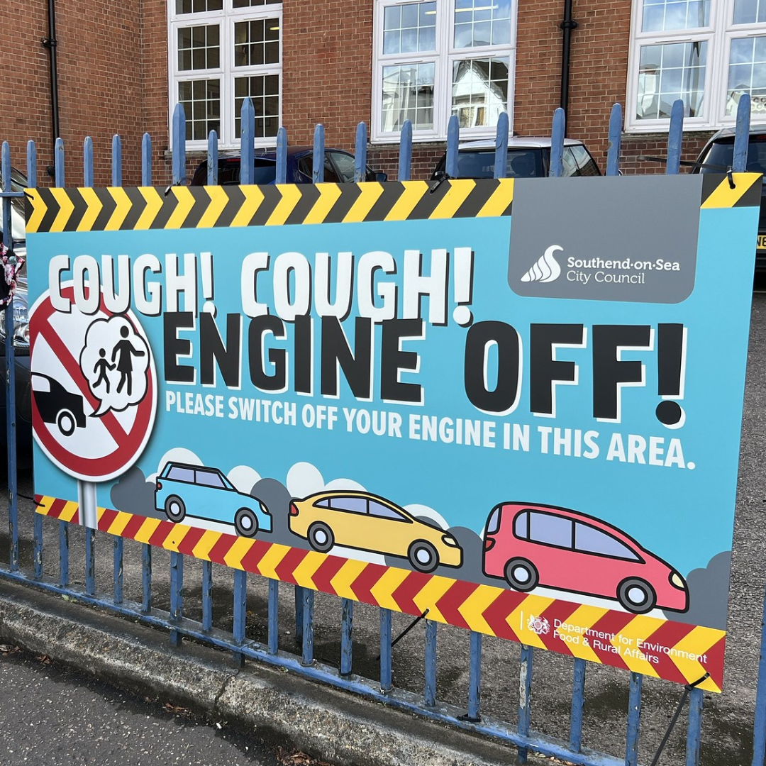 An Illustarted banner that is displayed along some metal railings outside of a school. It contains zigzag warning lines and some colourful cars. The text reads: "COUGH! COUGH! ENGINE OFF! Please switch off your engine in this area.