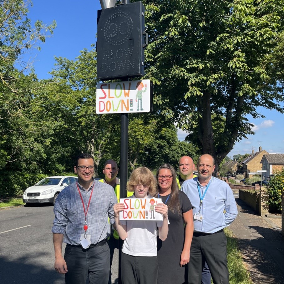 Photograph of Edward holding his sign underneath a lamp post on which the sign is displayed. Edward is surrounded by 5 adults.