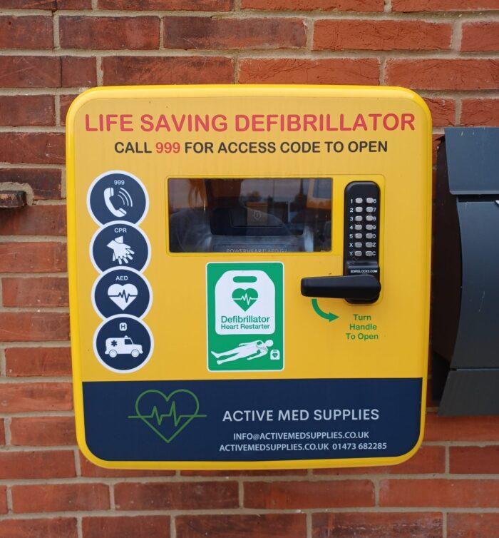 A yellow public difibrillator cabinet, mounted on a brick wall