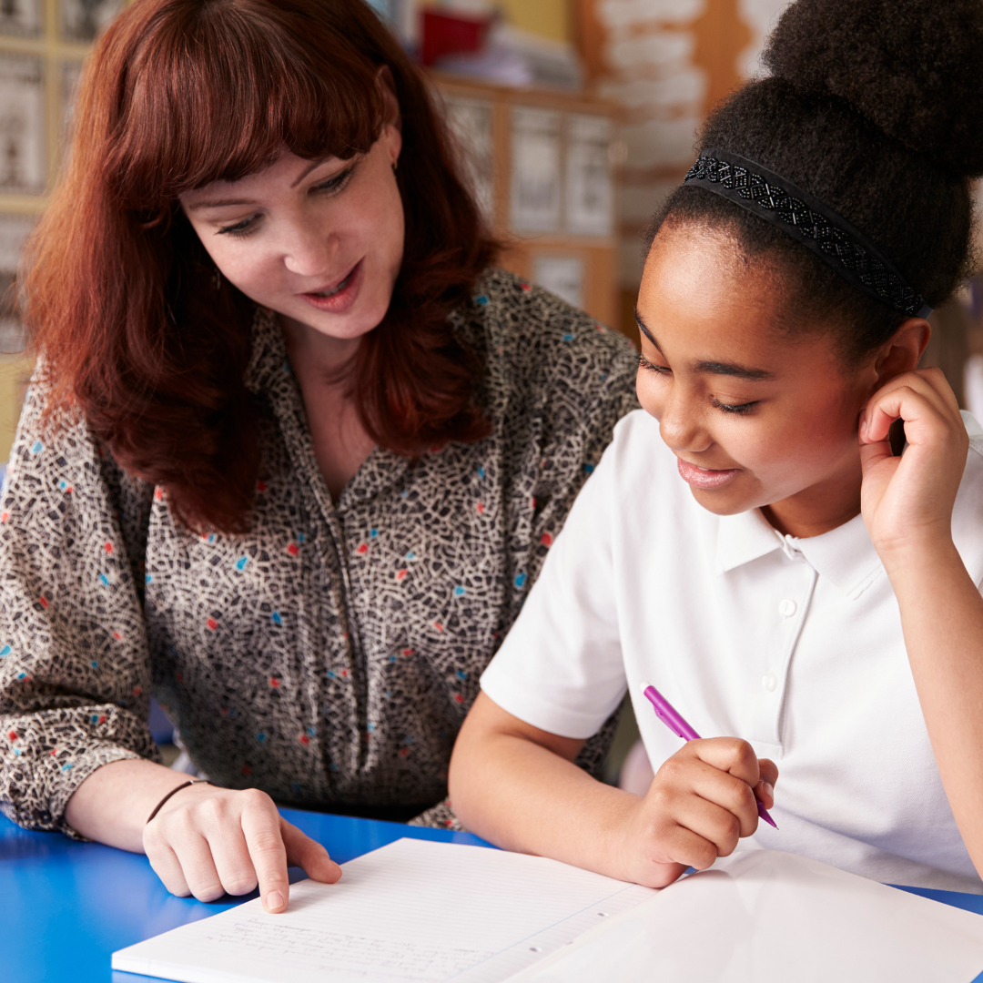 Photograph of a female teacher sat next to a female early teenage school student. The teacher points to something in a book in front of the student.