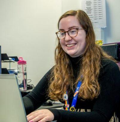 A woman with long, curly hair and glasses is smiling while working on a laptop. She is wearing a black turtleneck and a colorful lanyard with badges. There is a pink water bottle on the desk next to her, and the background shows an office setting