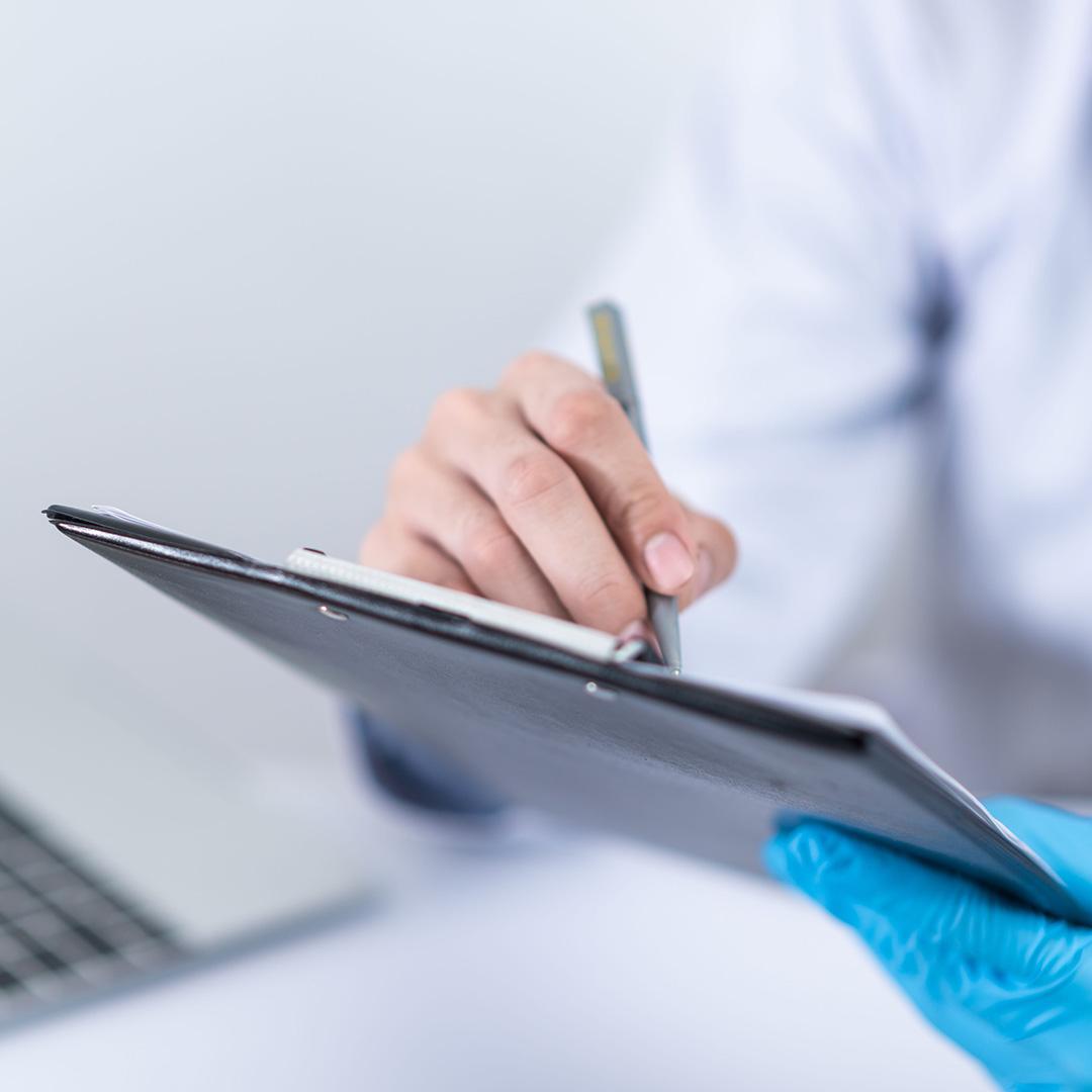 A hand with a stylus writing on a tablet. The person is wearing a white doctor&#039;s coat and has a blue disposable glove on their left hand, which is holding the tablet.