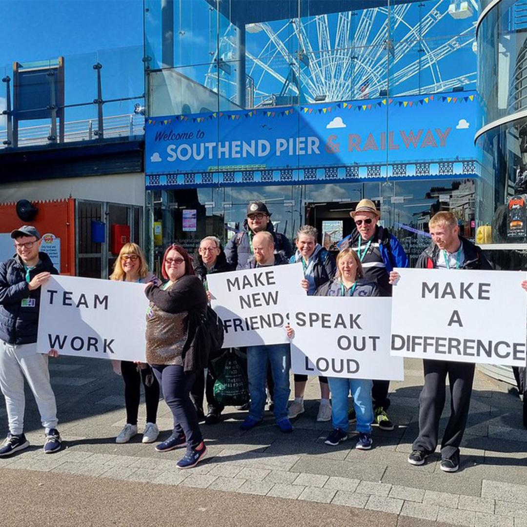 A group of people standing in front of the "Southend Pier & Railway" entrance, holding large white signs with bold black text.