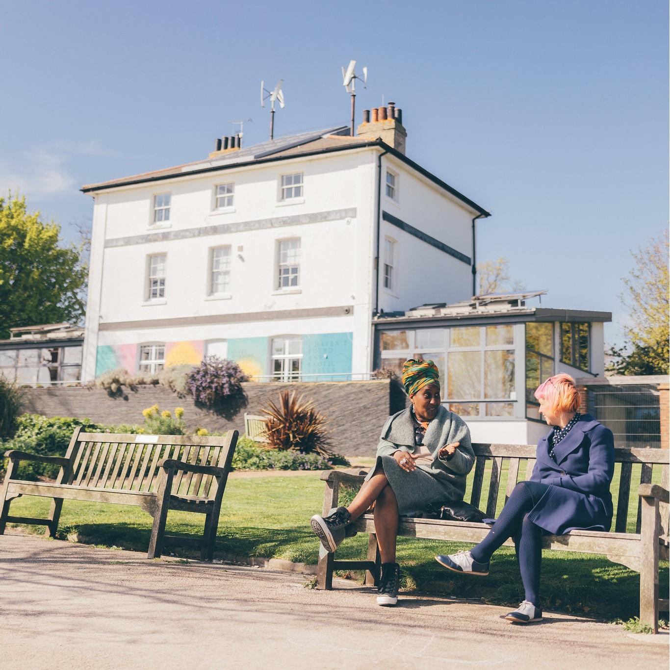 Two women sitting on a bench chatting in Chalkwell Park. The sky is blue and the building in the park can be seen in the background