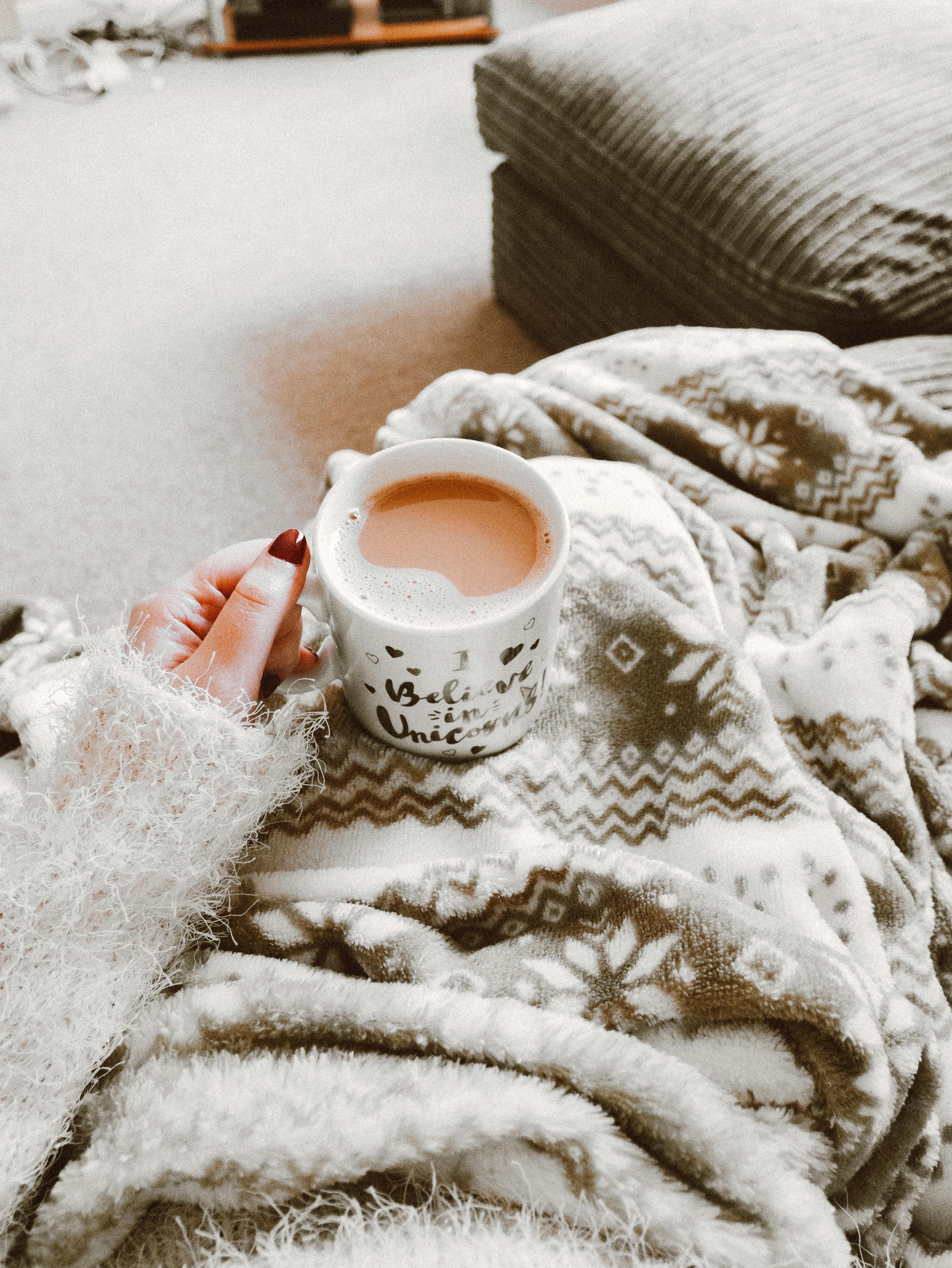 Persons hand holding a mug of coffee on their blanketed lap.