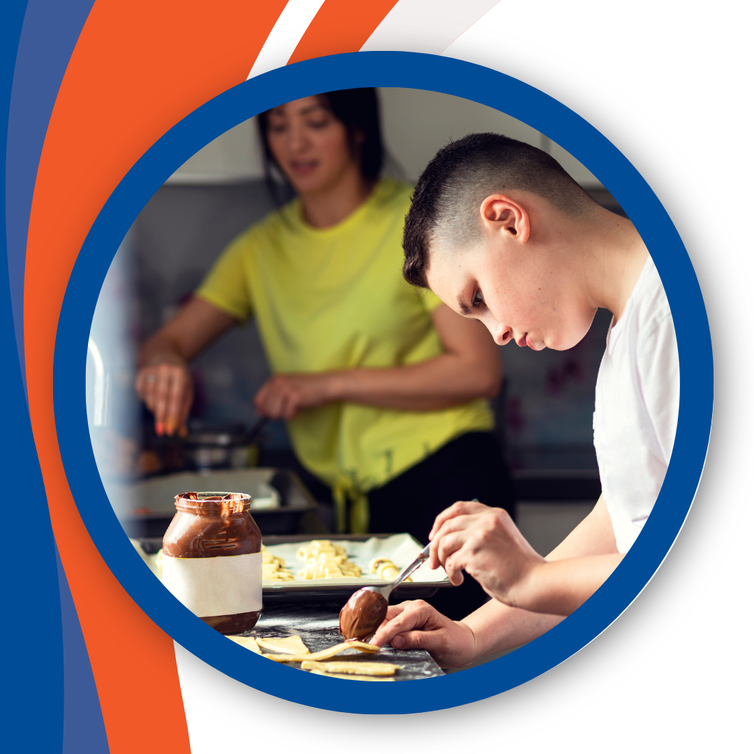 Photograph of a young person cutting some food up on a chopping board. Another young person is in the background stirring a pot on a hob